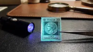 Francobollo Italia 20 lire Siracusana su superficie scura con luce UV e pinzette per verifica fluorescenza