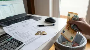 Mano che getta banconote in un cestino accanto a bollette, calcolatrice e computer con foglio di calcolo