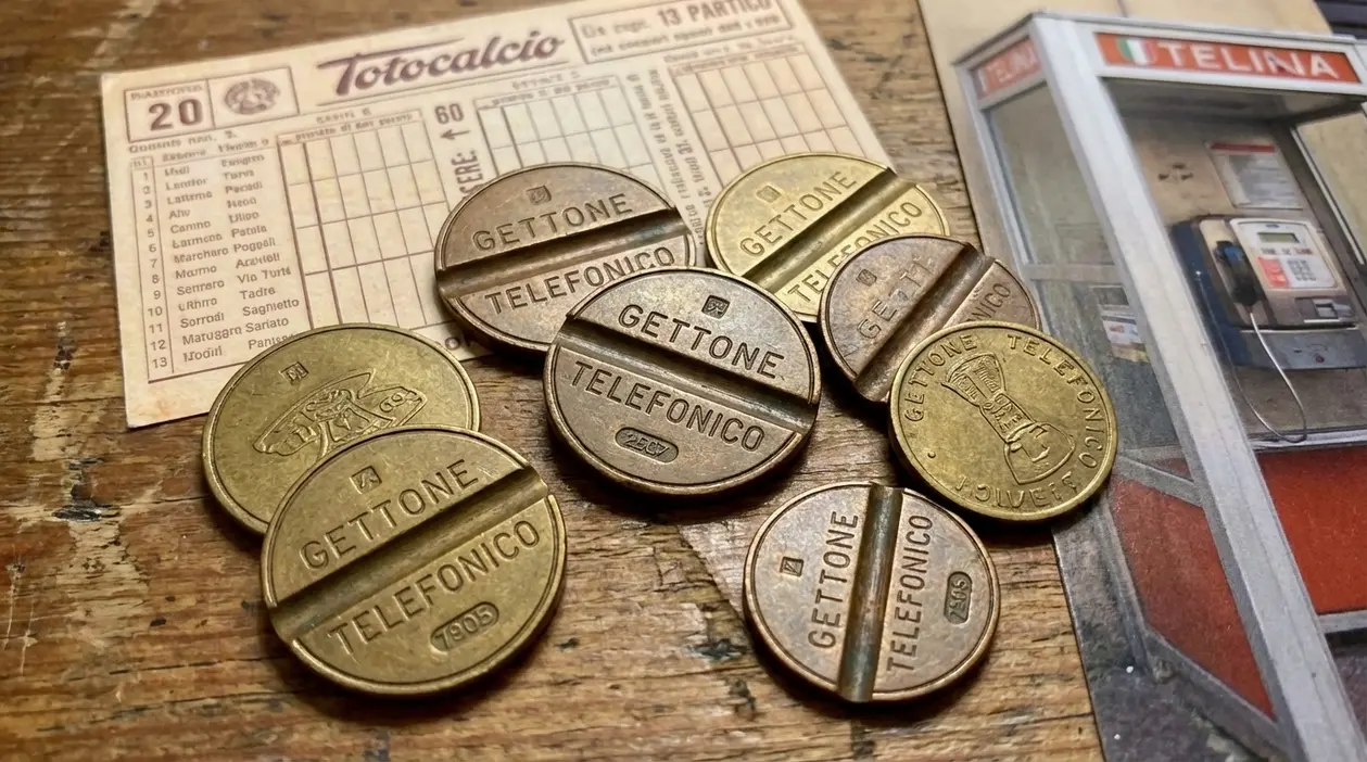 Gettoni telefonici d’epoca su un tavolo accanto a una schedina del Totocalcio e a una cabina telefonica