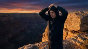 Persona in felpa scura in cima a un canyon al tramonto, accanto a una roccia con simboli zodiacali