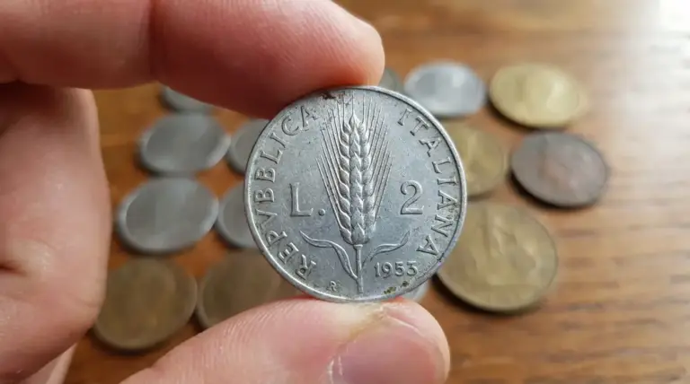 Primo piano di una moneta da 2 lire Spiga 1953 tenuta tra le dita, con altre monete sullo sfondo