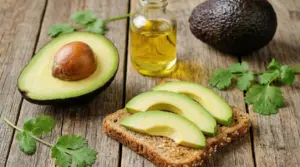 Fette di avocado su pane integrale con avocado intero, mezzo avocado e olio su sfondo di legno