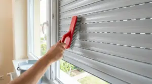 Una persona pulisce una tapparella con una spazzola a setole rigide dall'interno della finestra.