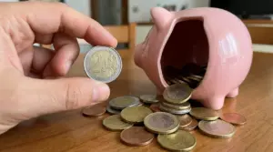 Mano che mostra una moneta da 2 euro vicino a un salvadanaio rosa rovesciato con monete sparse sul tavolo