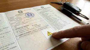 Dettaglio del libretto di circolazione con un dito che indica un simbolo di avvertimento; chiavi e penna sul tavolo