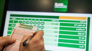 Mani che compilano una schedina del SuperEnalotto davanti a uno schermo con tabella di quote e premi