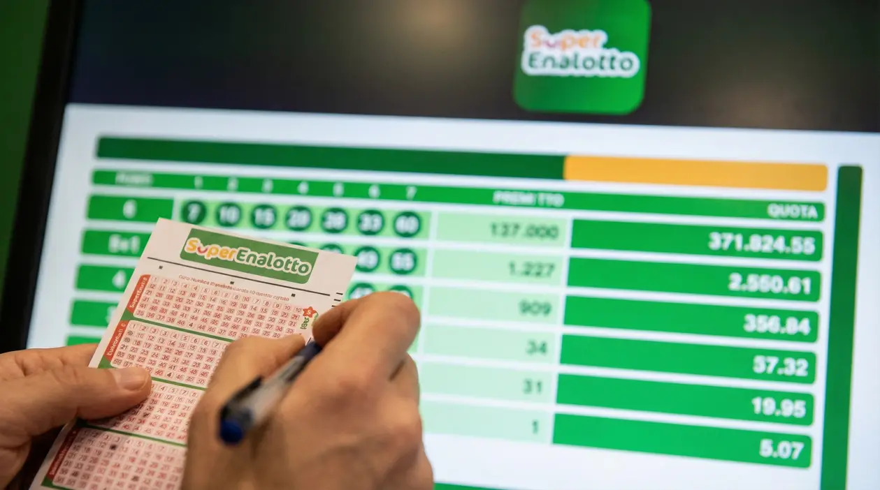 Mani che compilano una schedina del SuperEnalotto davanti a uno schermo con tabella di quote e premi