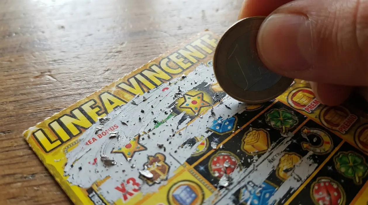 Mano che gratta un biglietto gratta e vinci con una moneta, cercando un simbolo nascosto sul tagliando